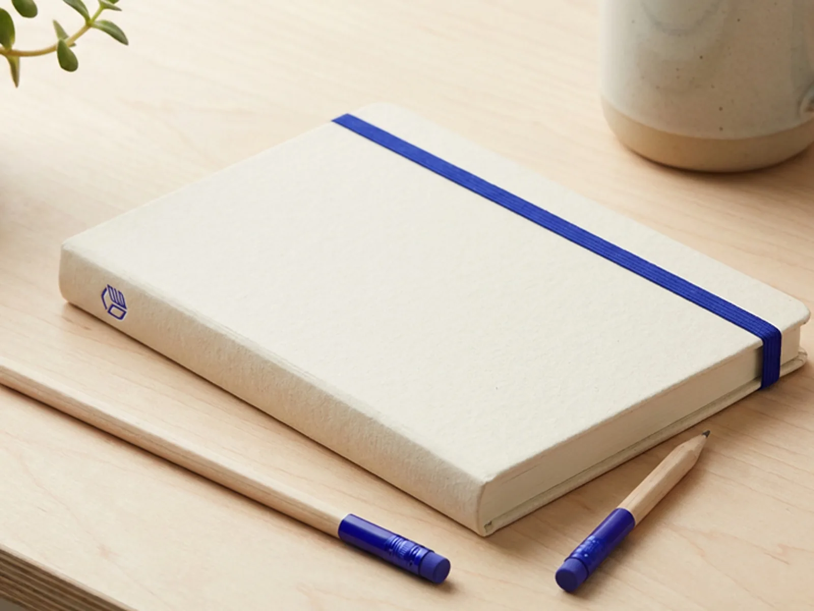 Libreta artesanal de BlunBlun cerrada con elástico azul, sobre un escritorio de madera junto a dos lápices.